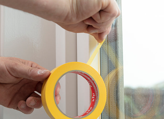 Afplaktape wordt aangebracht langs het glas van een houten buitenkozijn ter voorbereiding op schilderwerk - media 1
